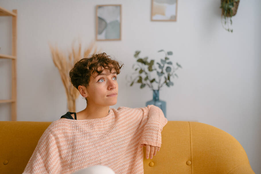 A woman in a piink sweater sits on the couch and gazes off with a hopeful expression.