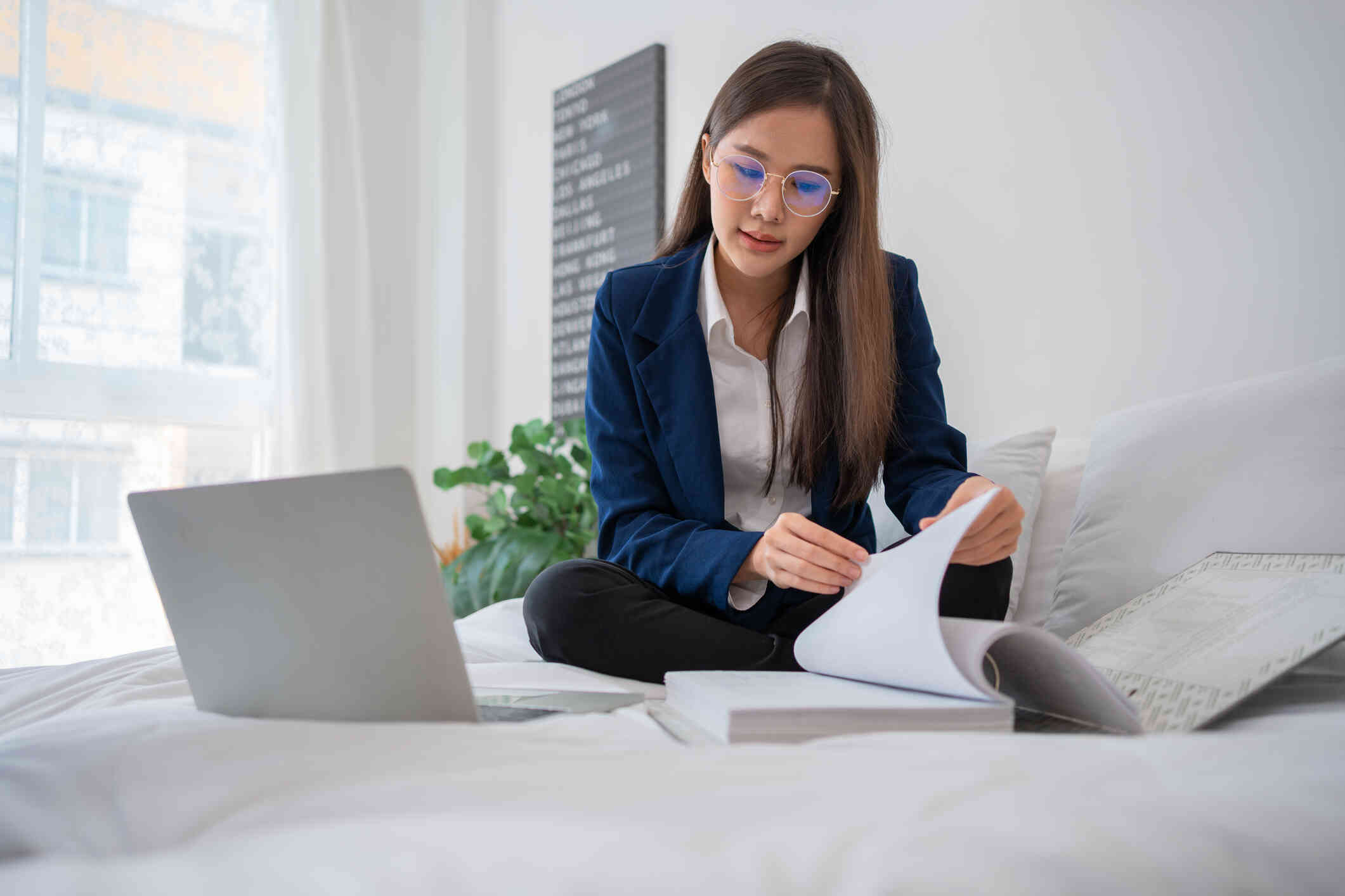 A woman sits on her bed and with her laptop open next to her as she flips through some some pages of a large binder.